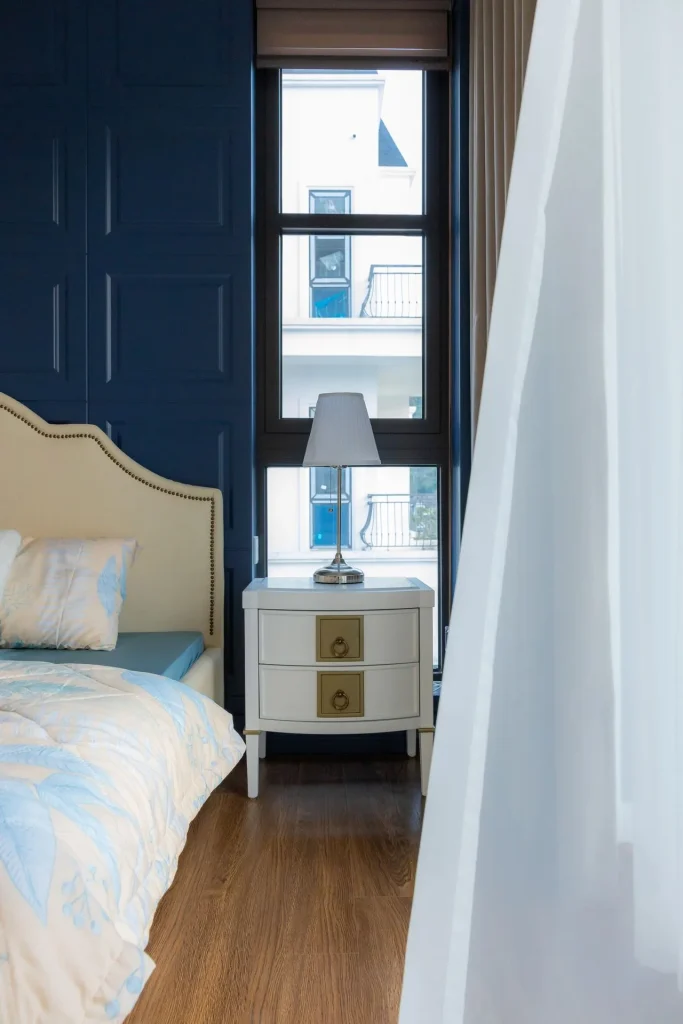 Quarto com cabeceira clara, parede azul e piso de madeira iluminado pela janela.