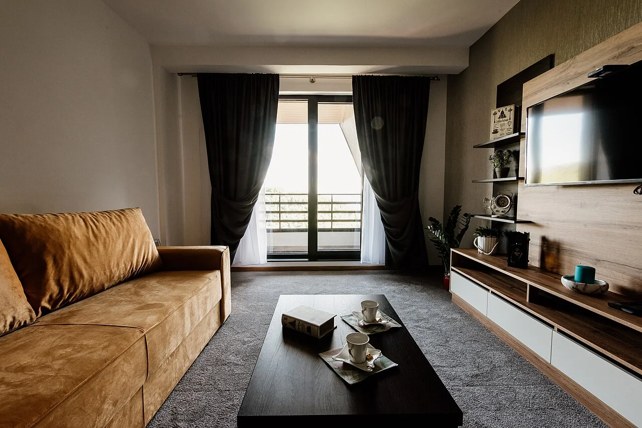 Sala de apartamento com sofá claro, móveis de madeira e janela ampla para a área externa.